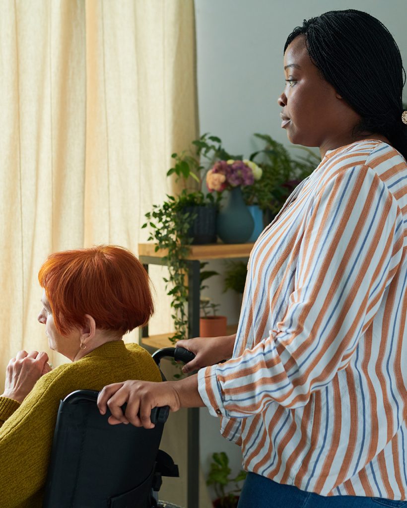 Senior woman with disability on wheelchair looking at window with African caregiver standing behind her in room